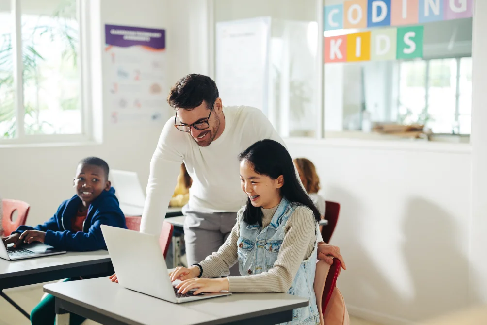 A teacher helping a student at a laptop in a bright classroom